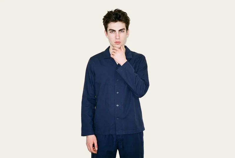 A 20-year-old Slavic man wears a navy cotton pyjama set in an upper-body front view. His wrist is held at his jaw, and he has a neutral expression against an alabaster flat-color backdrop.