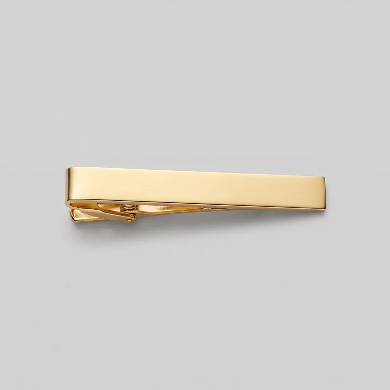 A gold tie bar is shown alone from a slightly low angled side view. Its smooth, elongated rectangular front bar has softly rounded corners and slightly beveled edges, while the underside reveals the hinged alligator-style mechanism, pivot pin, coil spring, lower gripping jaw, and small lever beneath the front plate.