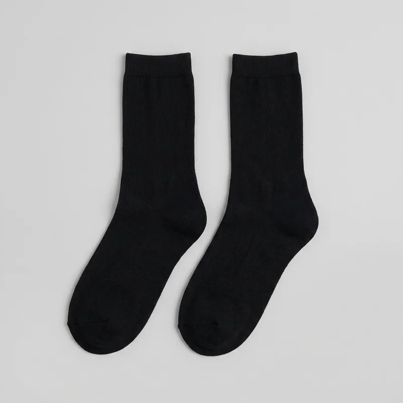 A complete pair of black close-fitting crew-length socks lies flat against a backdrop. The socks have straight tubular legs, ribbed cuffs, shaped heels, rounded closed toes, and fine vertical ribbing across the legs and feet. Seam shaping is visible at the heels and toes, with no visible logos, prints, patches, embroidery, or embellishments.
