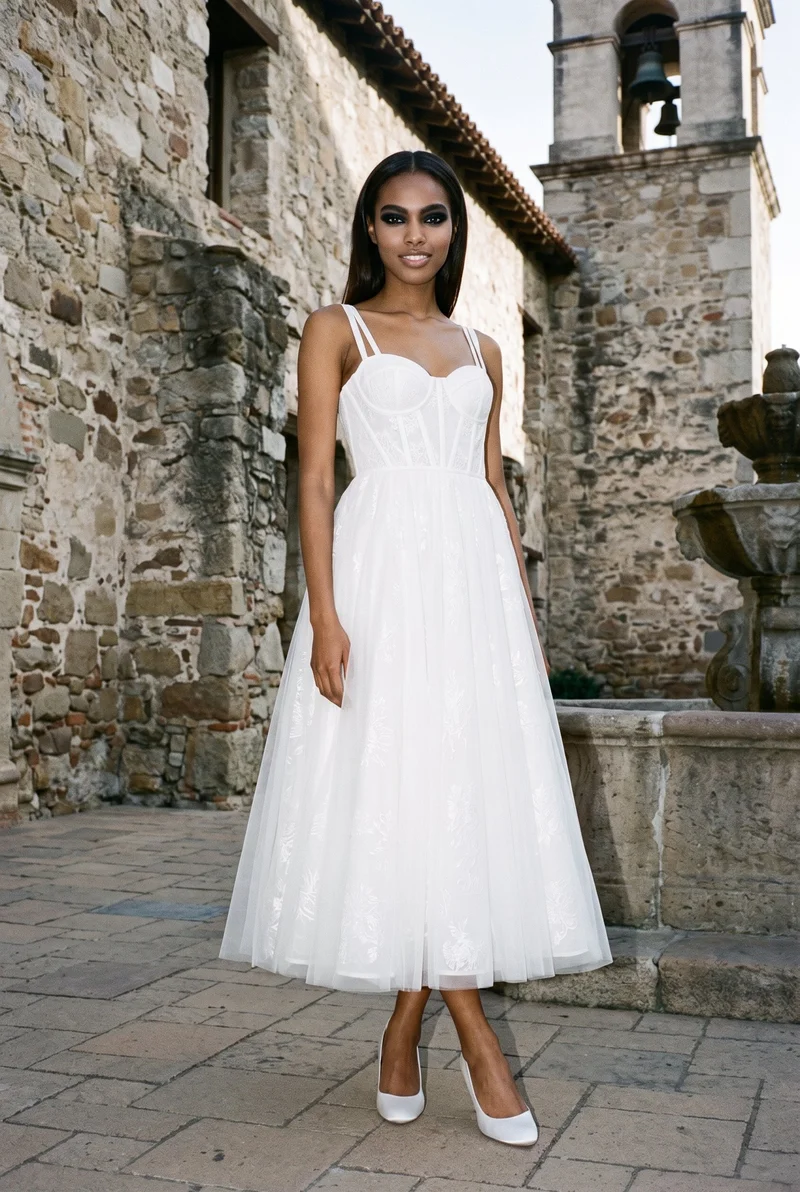 A 20-year-old African woman models a white A-line wedding dress in a full-body angled-front view, standing with her ankles crossed and smiling. The Mission Courtyard Bell Fountain appears as the backdrop.
