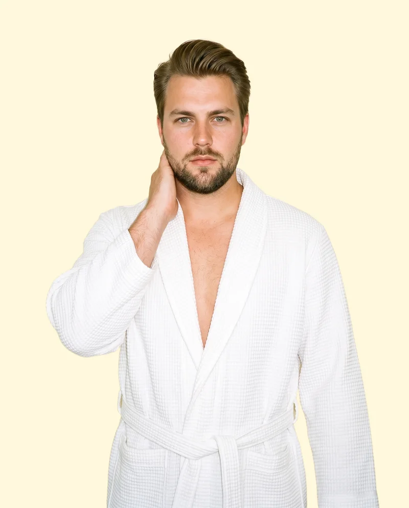 A 29-year-old Caucasian man is shown from the upper body in a front-facing view, wearing a white waffle robe with one hand on his neck and a natural facial expression. The image uses a flat buttercream backdrop.