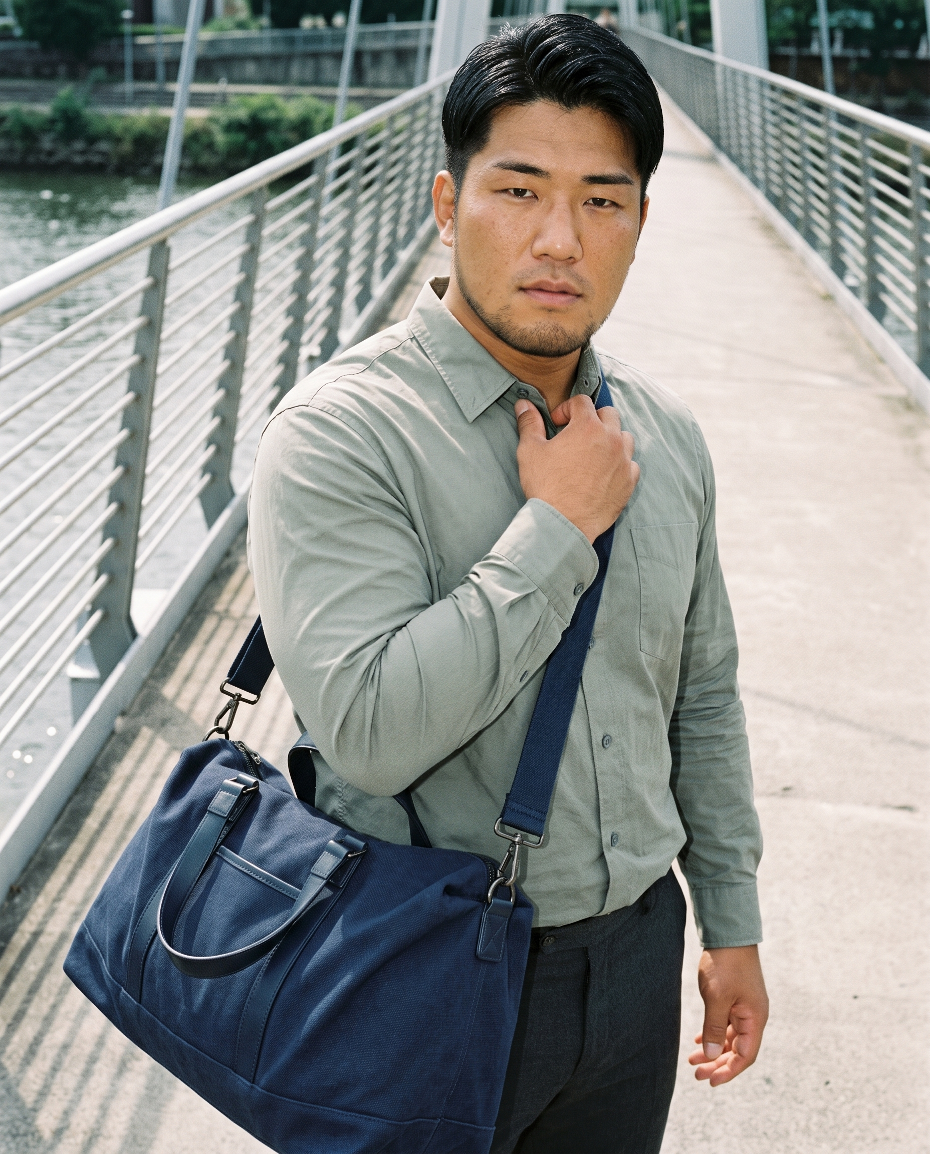 A 31-year-old Japanese man is shown from the upper body in a front view, wearing or carrying a navy canvas weekend bag. One hand rests at his collarbone, and he has a soft facial expression. A bridge pedestrian backdrop is visible behind him.