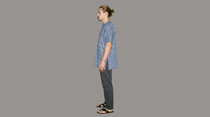 A 20-year-old Caucasian man wears a printed kaftan cover-up in an environmental side-view composition. He is shown with a waist twist and a serious expression against a flat studio-grey backdrop.