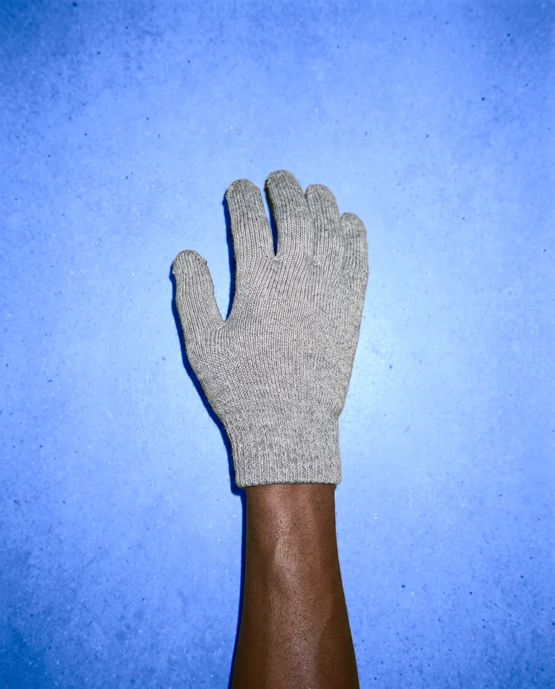 A man's hand and wrist wearing a grey knit glove, shown from above with the hand at rest. The denim blue studio-wall backdrop frames the isolated hand and glove.