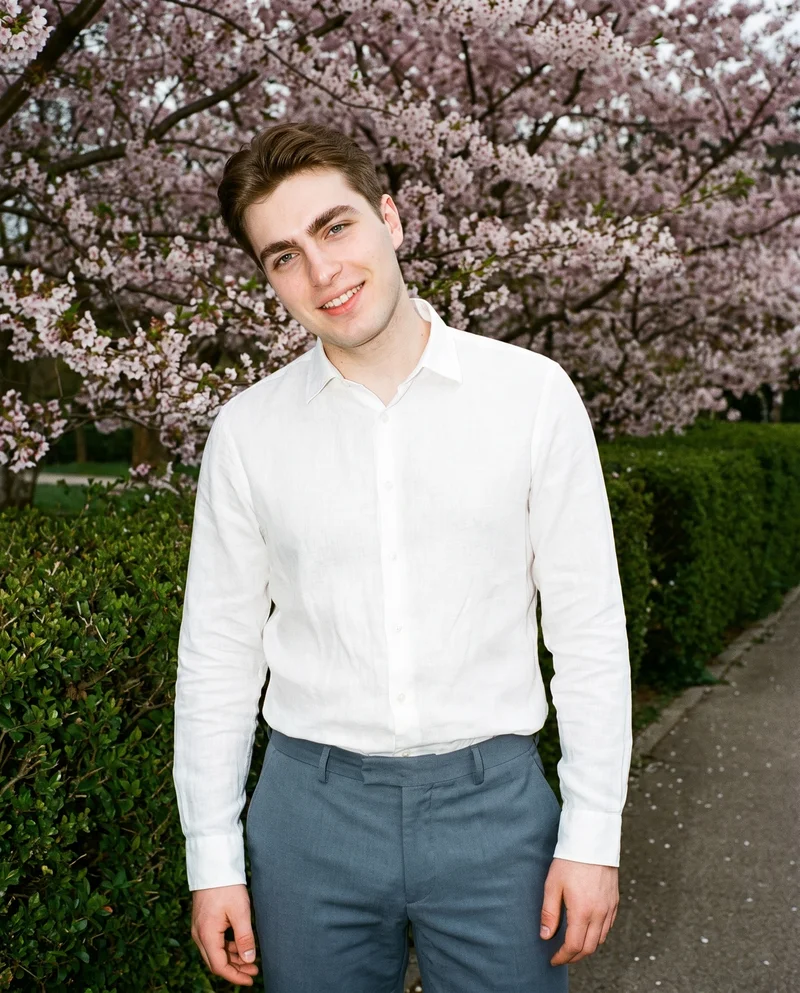 A 20-year-old Slavic man is shown from the front in an upper-body view wearing a white linen shirt. He is smiling with a shoulder-drop pose, against a Cherry Blossom Park backdrop.