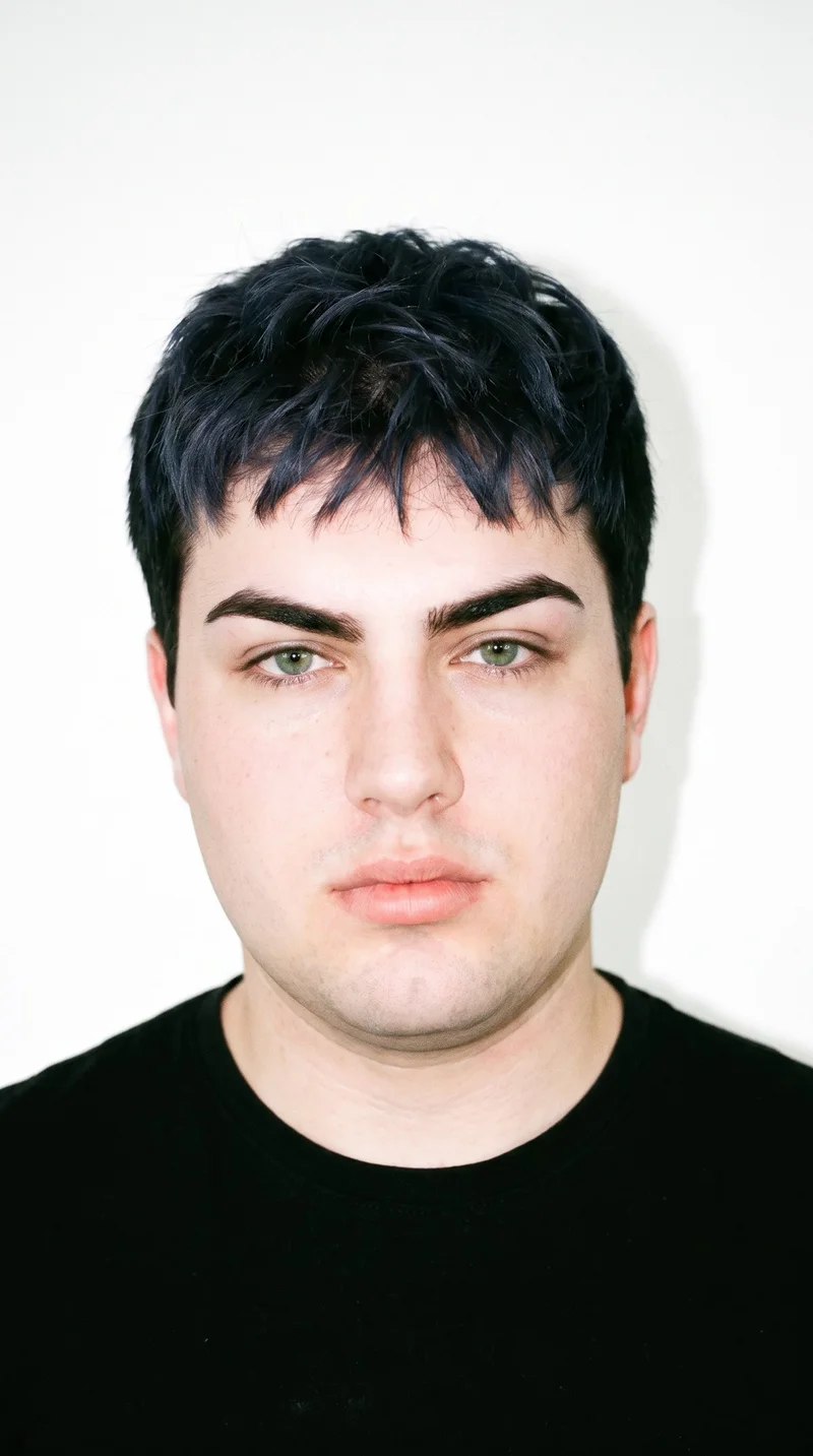 An 18-year-old Caucasian male with pale skin and a peach undertone. His face has sharp, angular features, pronounced cheekbones, and a defined jaw. He has thick eyebrows, green eyes, and full lips. His black hair is styled in a short textured crop with layered texture on top and closely cut sides. He is clean-shaven, with a smooth face.