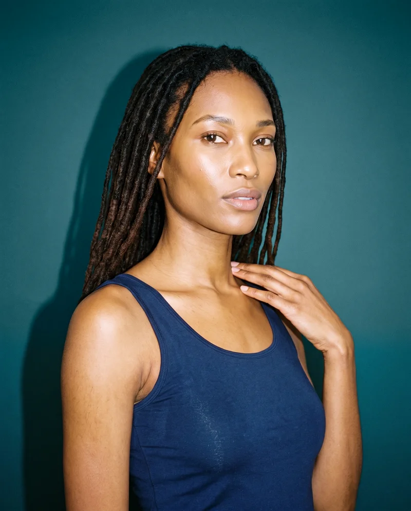 A 26-year-old African woman is shown in a bust-length front three-quarter view wearing a navy training tank. One hand rests at her collarbone, and she has a natural facial expression against a petrol studio-wall backdrop.