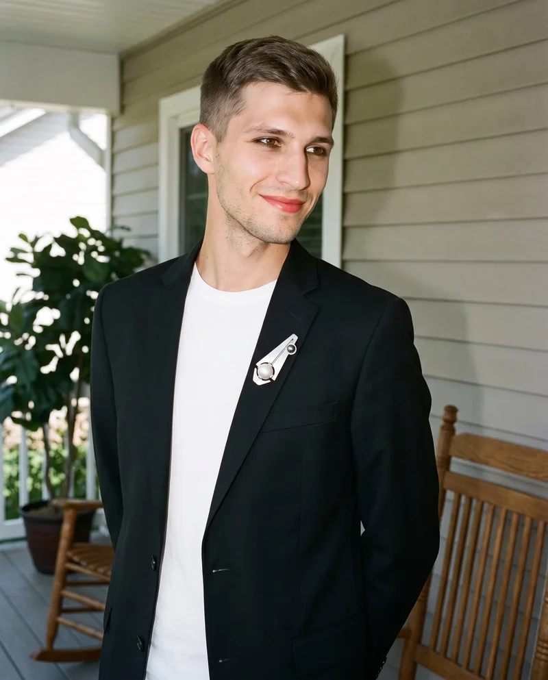 A 26-year-old Slavic man is shown from the upper body at an angled front view, smiling with his hands behind his back and wearing a men's pearl brooch. A front porch serves as the backdrop.