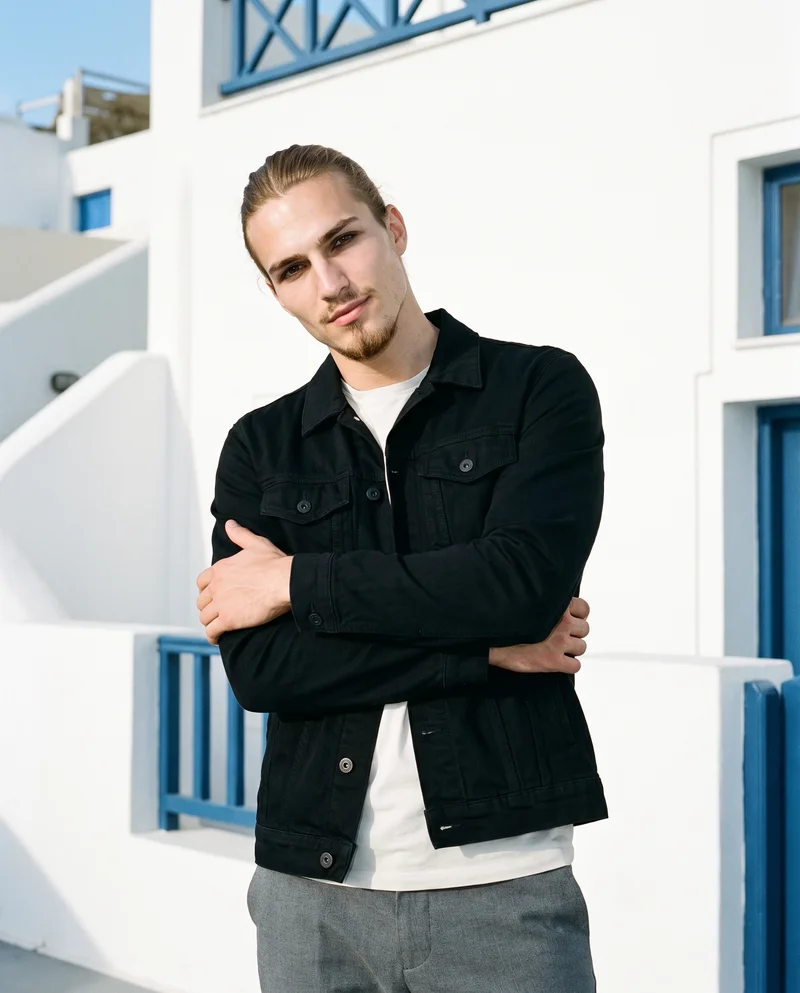 A 20-year-old Caucasian man is shown from the upper body at a front three-quarter angle, wearing a black denim jacket. He poses with his arms crossed in a self-hug stance and has a confident facial expression. A Greek Villa White Blue backdrop is behind him.
