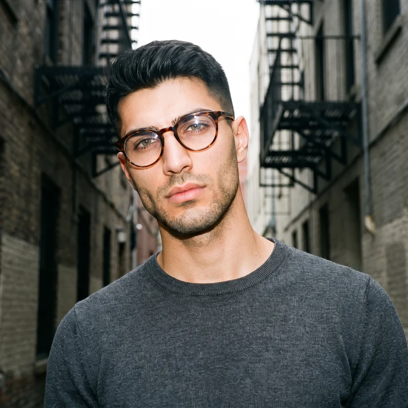 A 22-year-old Turkish man wears round blue-light glasses in a bust-framed front three-quarter view. His head is tilted with a neutral expression, against a city alleyway backdrop.