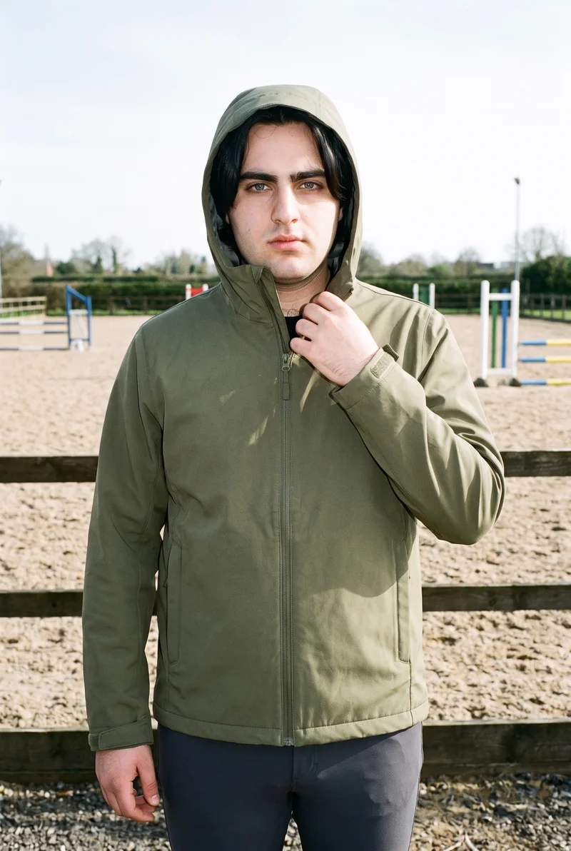 A 23-year-old Caucasian man is shown from the upper body in a front view wearing a green waterproof jacket. One hand rests at his collarbone, and he has a neutral expression. An equestrian arena forms the backdrop.