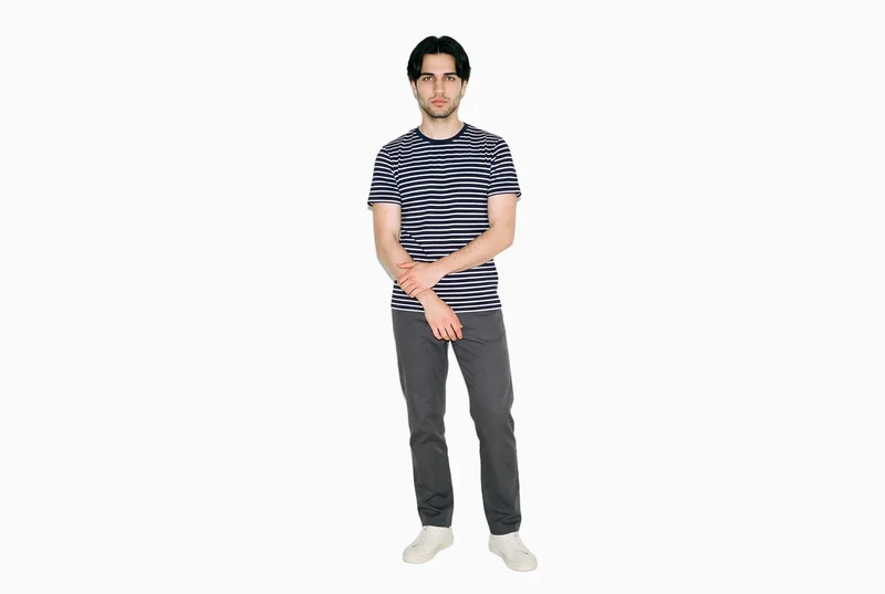 An 18-year-old Slavic male model wears a men's striped cotton T-shirt in a front-facing environmental composition. His arms are held low and his expression is natural, with a flat seasalt-colored backdrop behind him.
