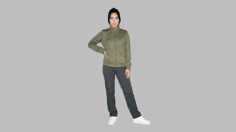 A 25-year-old Hispanic woman wears an olive tracksuit top against a dove grey backdrop. She is shown in an environmental frame from a front three-quarter angle, standing with one foot forward and a confident expression.