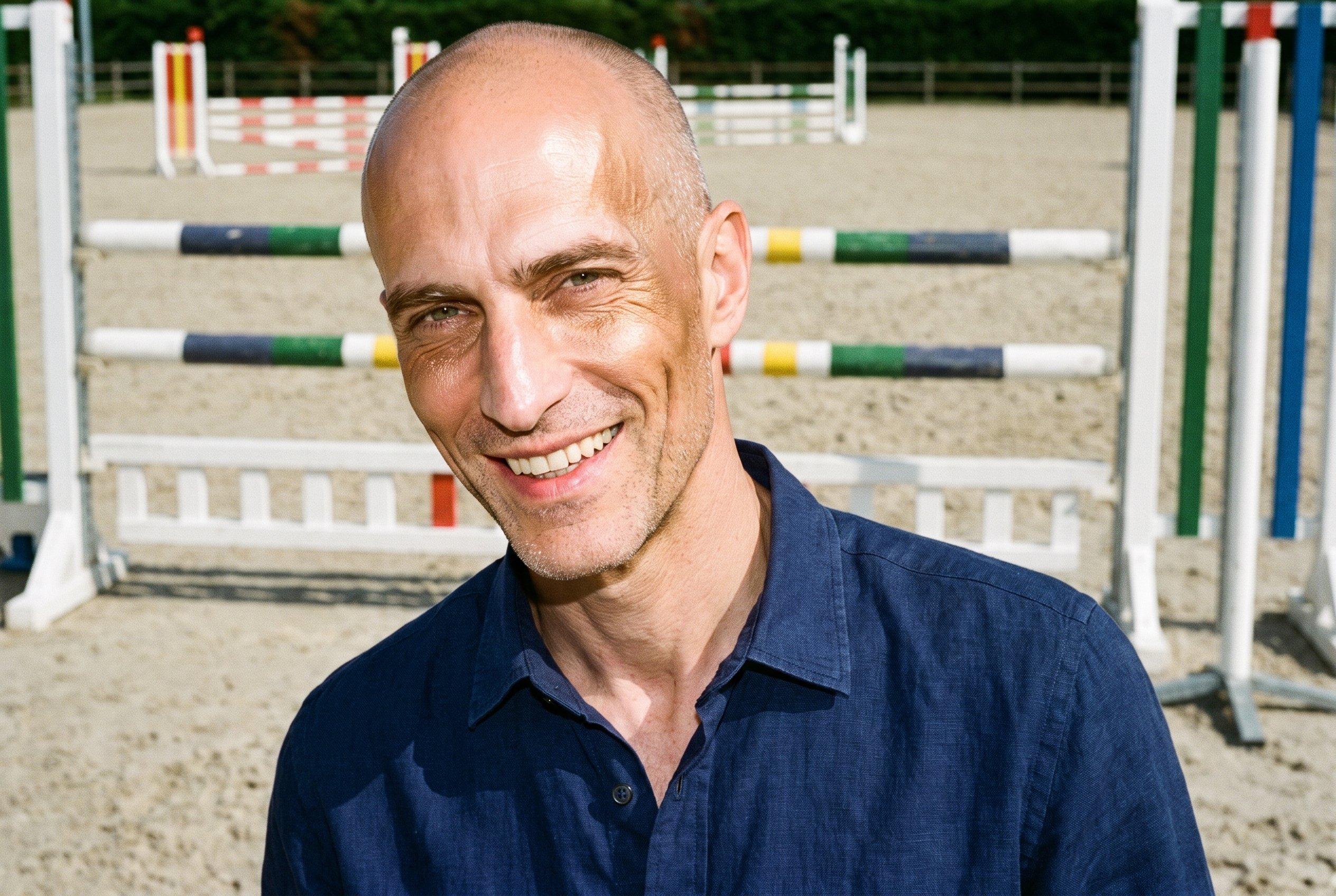 A 37-year-old Caucasian man is shown from the front in a bust composition, wearing a navy linen shirt. He tilts his head and grins against an equestrian arena backdrop.