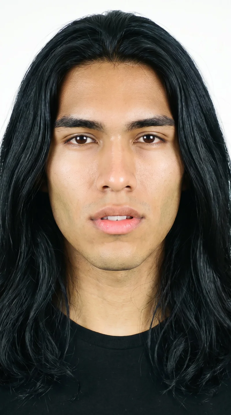 An 18-year-old Hispanic male with a medium olive skin tone and a warm, greenish-golden undertone. His face has a triangular structure, with a wide jaw that narrows toward the forehead and emphasizes the lower face. He has medium-thickness eyebrows, brown eyes, and full lips. His deep black hair is long, worn loose past the shoulders with natural volume. He is clean-shaven, with smooth skin across the whole face.