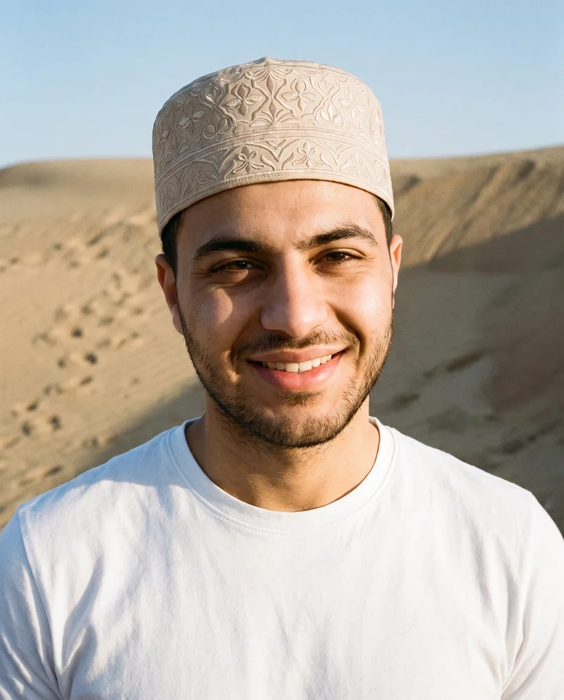 A 20-year-old Arab man is shown from the bust in a front view, wearing a beige embroidered kufi. His head is tilted with a natural expression, against a sand dunes and footprints backdrop.