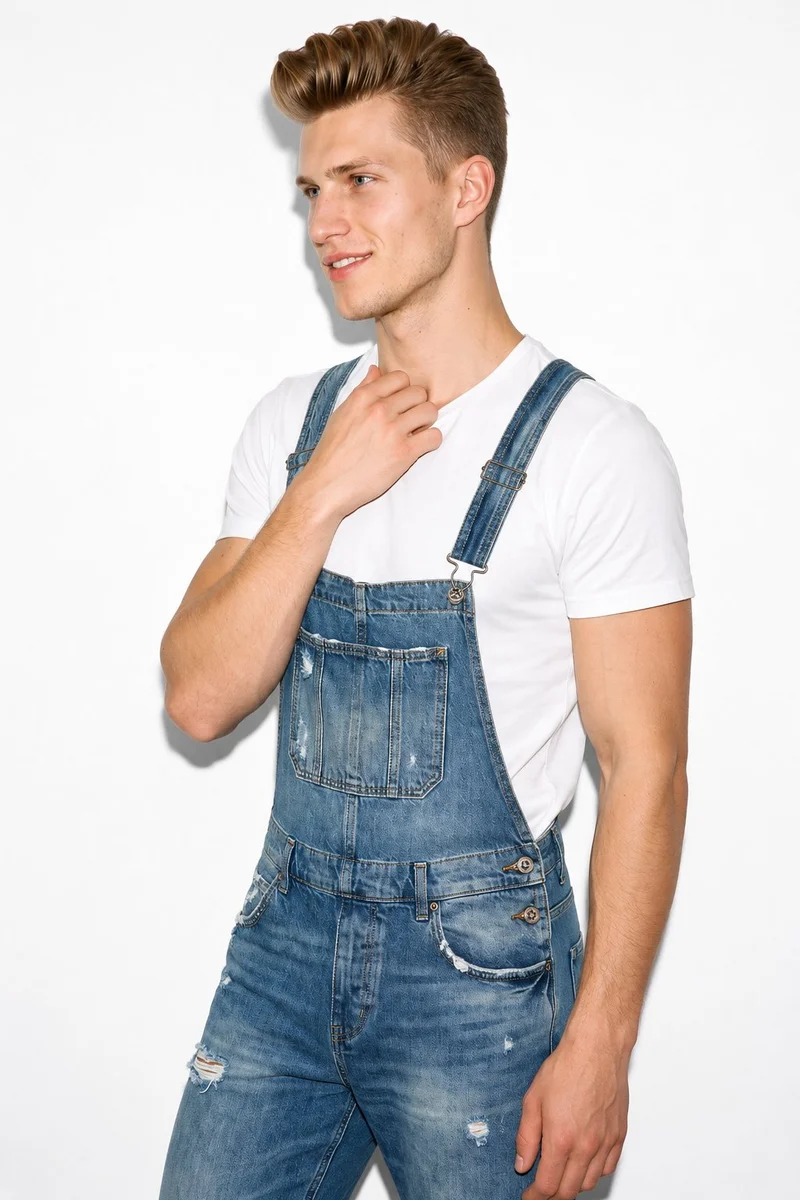 A 21-year-old Caucasian man wears distressed denim dungarees in an upper-body composition. He is shown from an angled front view with one hand at his collarbone and a natural facial expression, against a pure white studio backdrop.