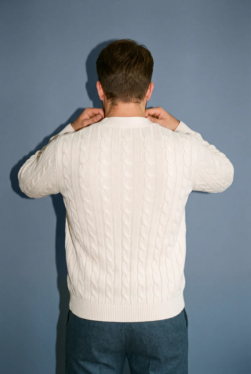 A 29-year-old Caucasian man is shown from the upper body in a back view, wearing a men's cream cable-knit cardigan with his hands on his neck. A slate grey studio-wall backdrop is visible.