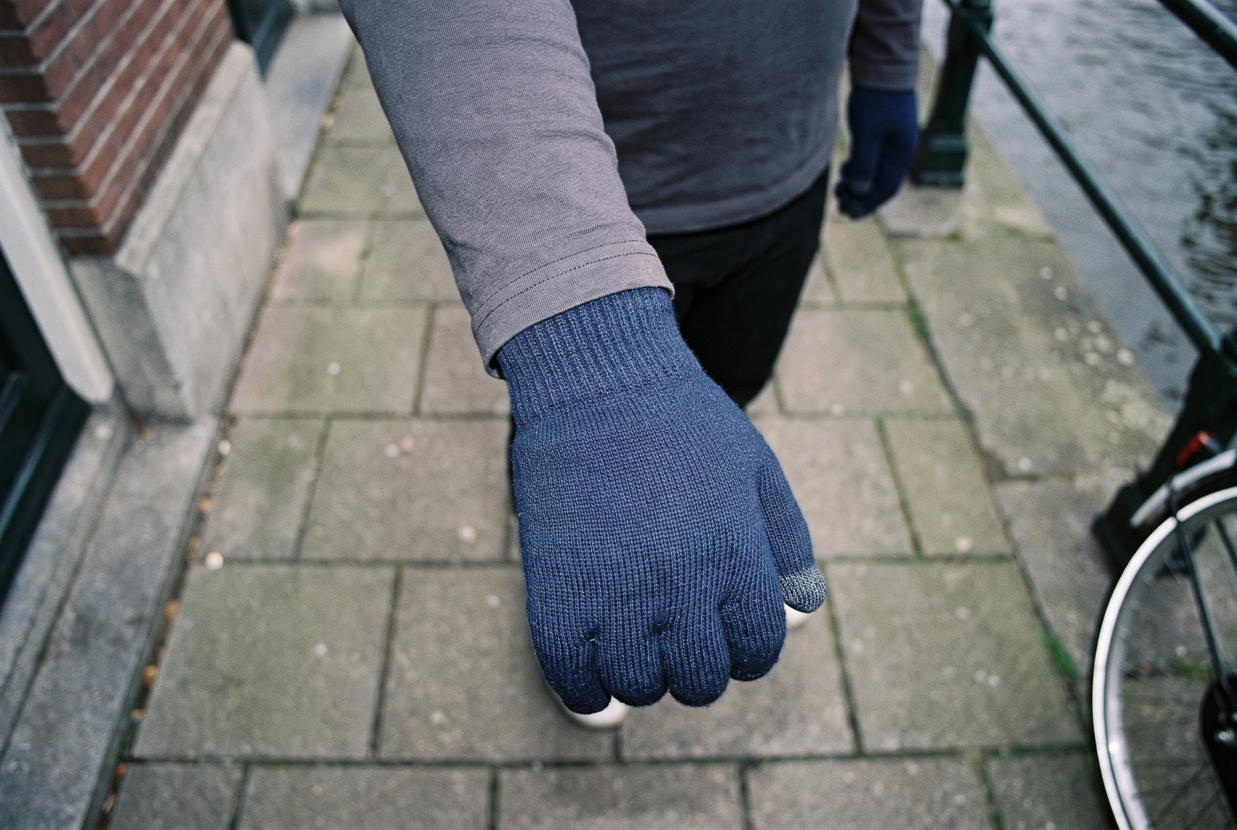 A close top view of a man's hand and wrist wearing navy touchscreen gloves, positioned with the knuckles facing forward. An Amsterdam canal house backdrop is visible.