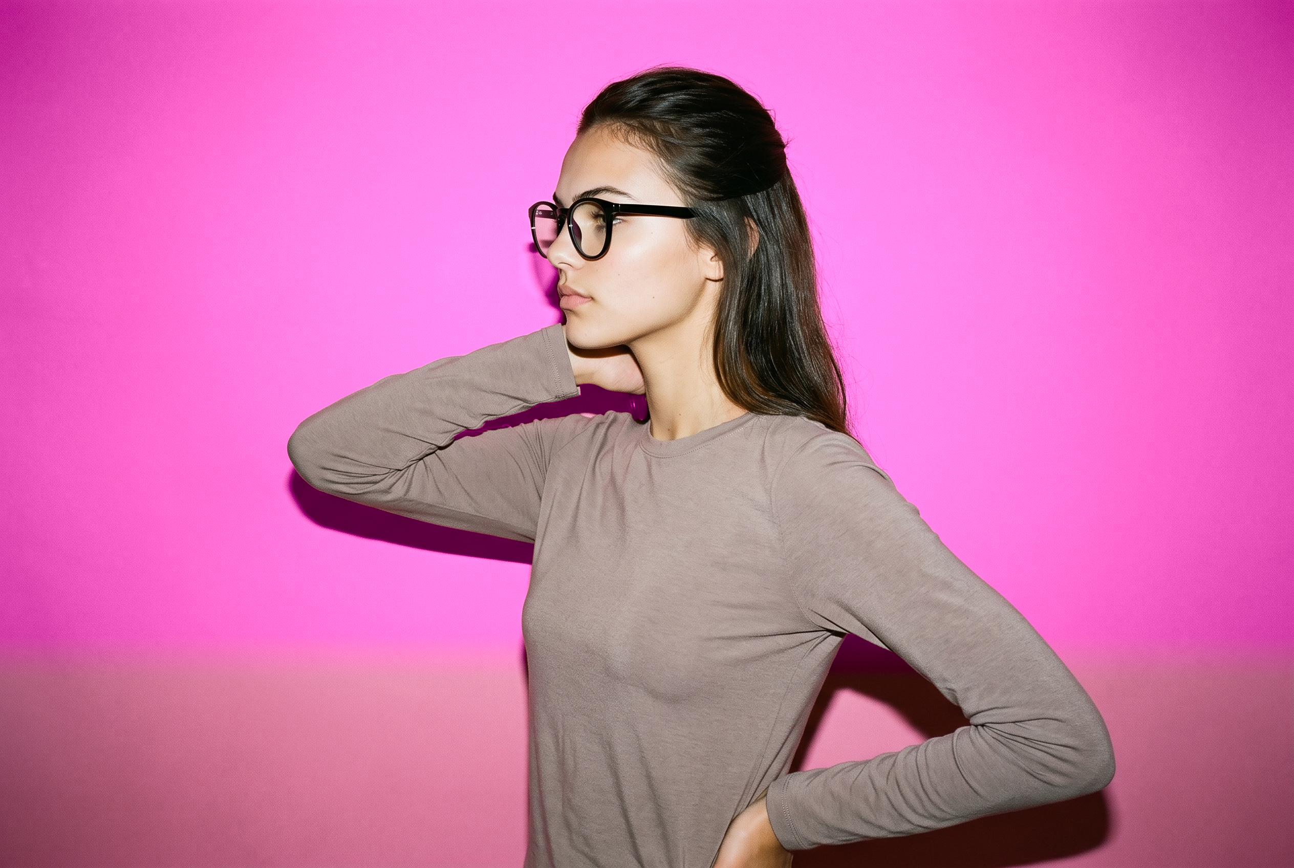 An 18-year-old Caucasian woman is shown in an upper-body side view wearing black blue-light glasses. Her hand is positioned behind her neck, and she has a soft facial expression against a rose-pink studio-wall backdrop.