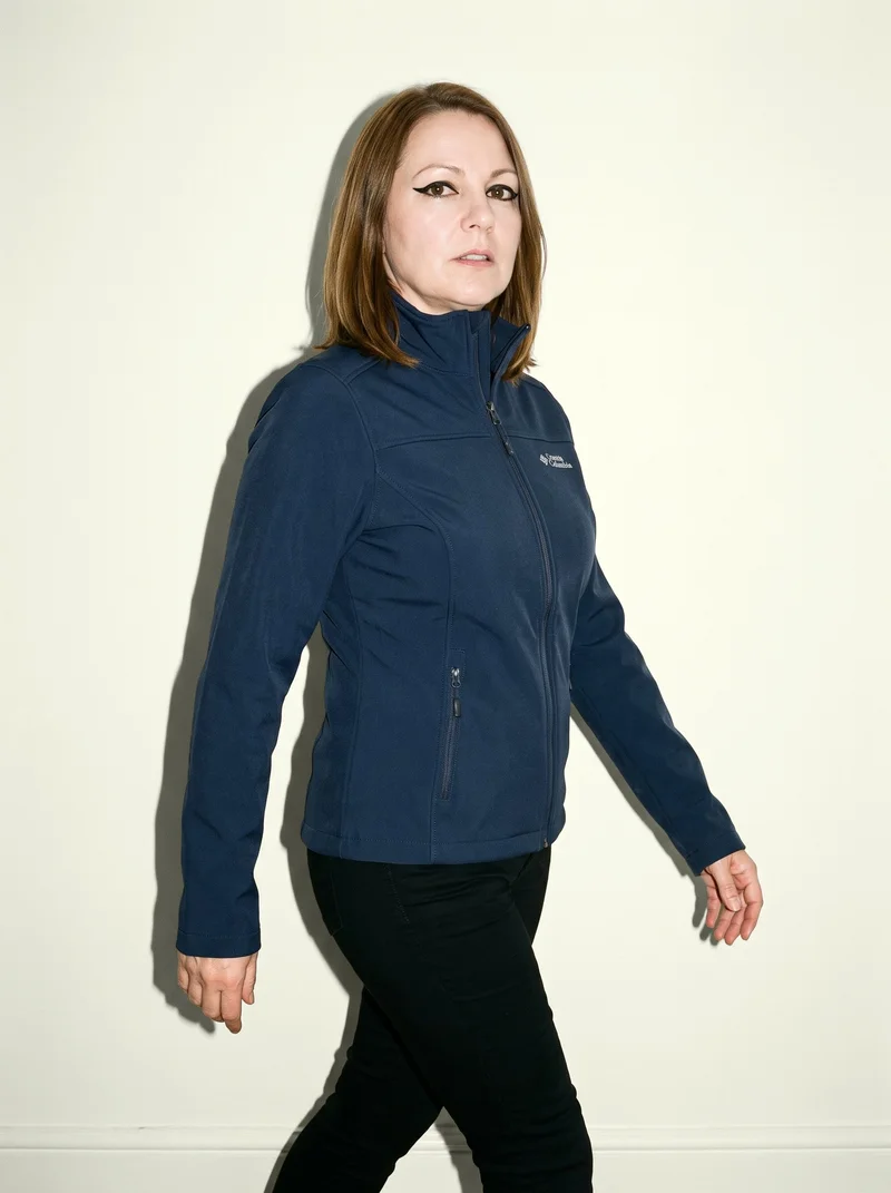 A 51-year-old Caucasian woman wears a women's navy softshell jacket in a three-quarter-length view, shown from an angled front perspective while walking with a neutral expression. An ivory lace studio-wall backdrop fills the background.
