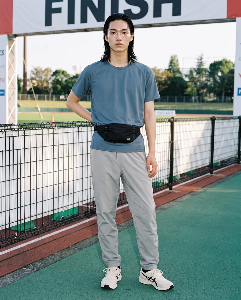 A 19-year-old Japanese man is shown full body from the front wearing a men's black belt bag. One hand rests on his lower back, and he has a natural expression. The backdrop is a marathon finish line.