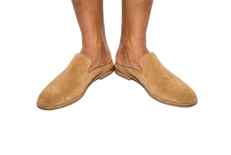 A pair of men's tan suede mules is shown in a side view with both feet framed and the toes pointing outward. The footwear is presented against a pure white backdrop.