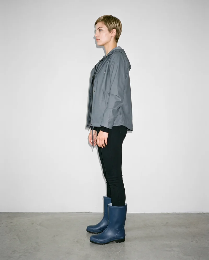 A 24-year-old Caucasian woman wears women's navy rubber rain boots in a full-body side view, standing with her hands in front and a fierce expression. A pearl-grey studio wall forms the backdrop.