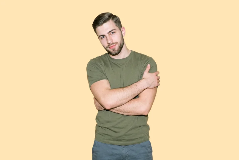 A 21-year-old Caucasian man is shown from the upper body in a front view, wearing an oversized olive T-shirt and holding himself in a self-hug pose. He has a confident facial expression against a flat peach backdrop.