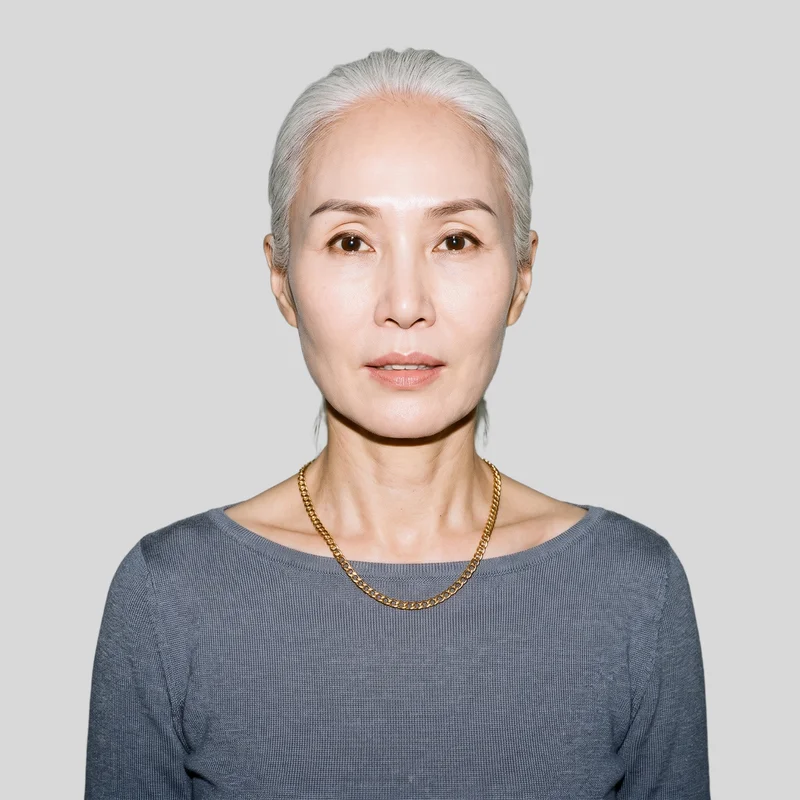 A 56-year-old Korean woman is shown from the front in a bust-length view with her shoulders square, wearing a women's gold chain necklace. The necklace is presented against a flat pearl-grey backdrop.