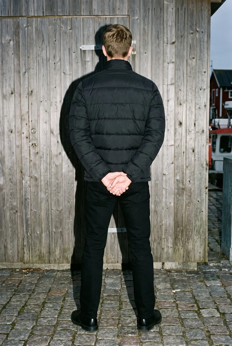 A 43-year-old Caucasian man wears a black down jacket in a full-body rear view, standing with his hands behind his back. A Cornwall fishing village forms the backdrop.