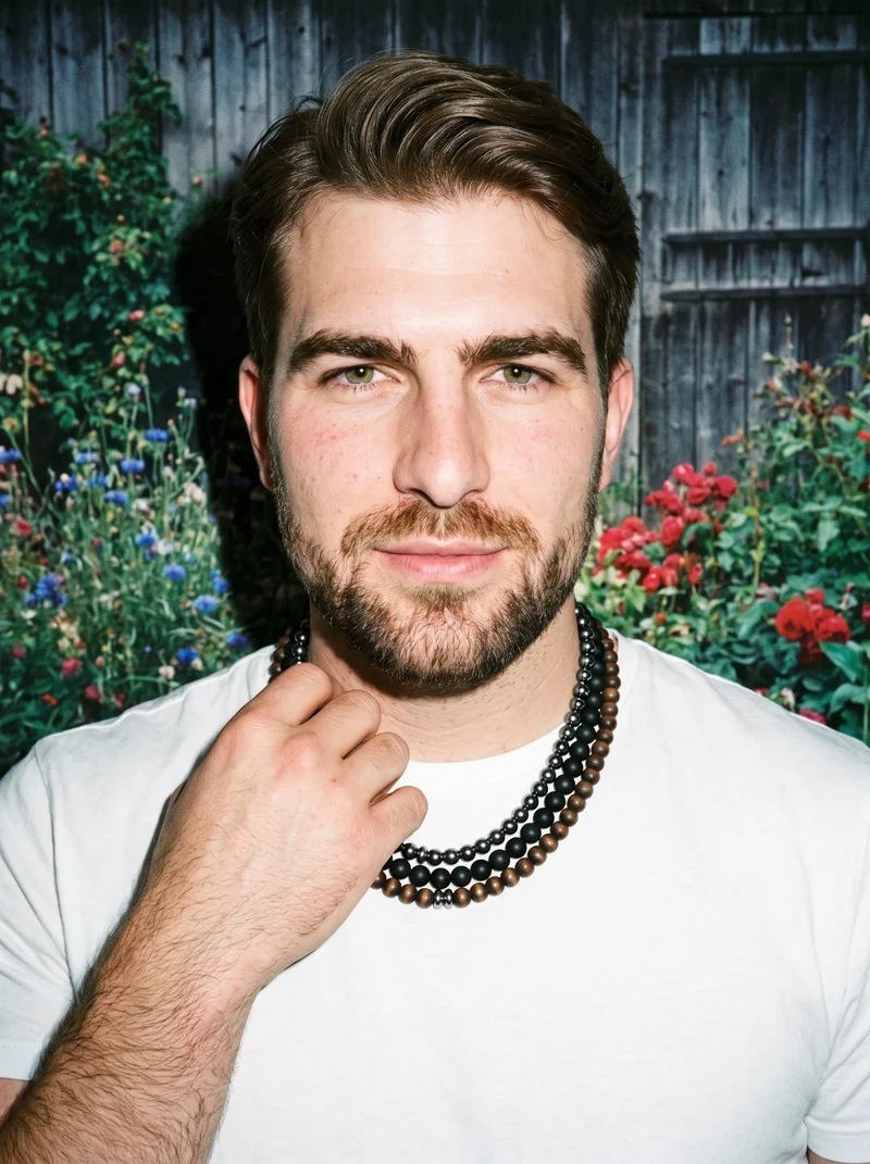 A 29-year-old Caucasian man is shown from the front in a bust-length view, smiling with one hand at his collarbone to display a men's layered beaded necklace. A cottage garden serves as the backdrop.