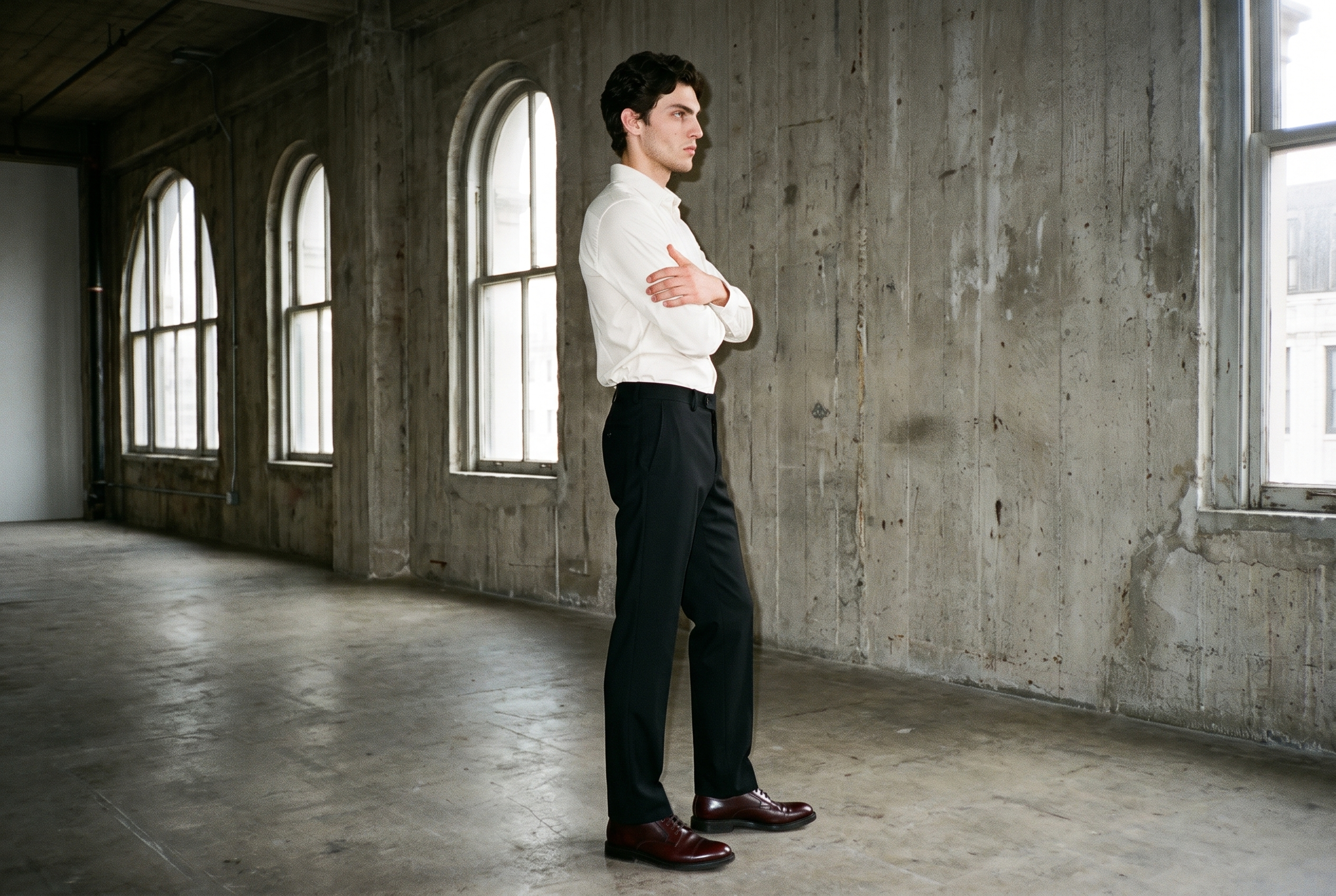 An 18-year-old Caucasian male model wears black tailored trousers in a full-body side view with a self-hug pose and natural expression. Massive loft windows with a golden backdrop appear behind him.