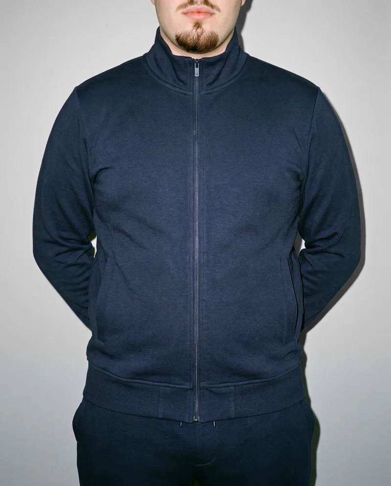 A male model wears a navy tracksuit in a front-facing torso crop, standing with his hands behind his back. A Silver Mist studio-wall backdrop fills the background.