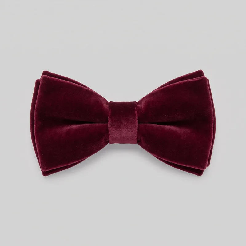 A complete burgundy velvet bow tie is shown standalone from the front. It has a symmetrical double-wing silhouette with flared rectangular ends, softly rounded outer corners, a pinched gathered center, and a vertical center wrap band. Layered folded edges are visible behind the front panels, giving the bow tie a slightly padded, dimensional structure.