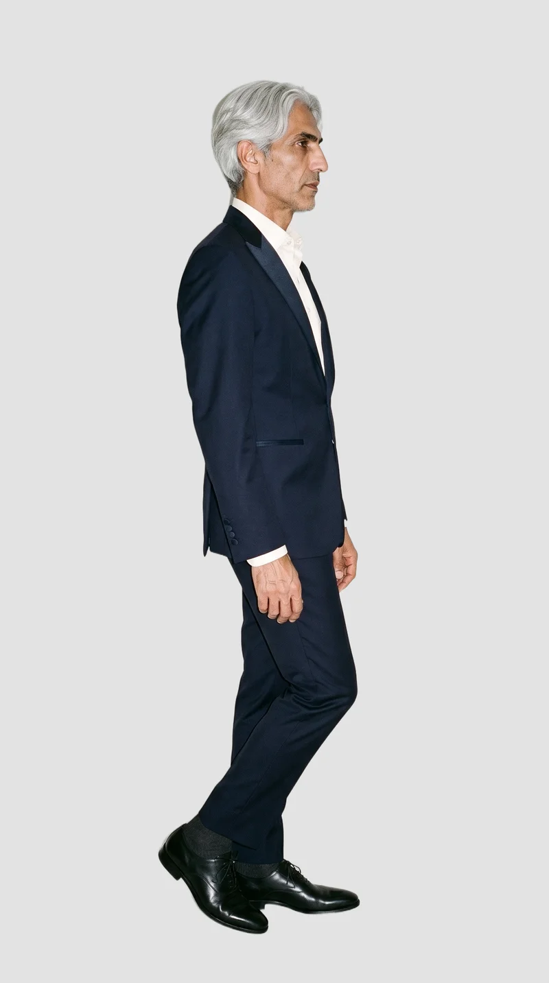 A 58-year-old Arab man wears a midnight blue tuxedo in a full-body side view, standing with his ankles crossed and a neutral expression. A flat fog grey backdrop fills the background.
