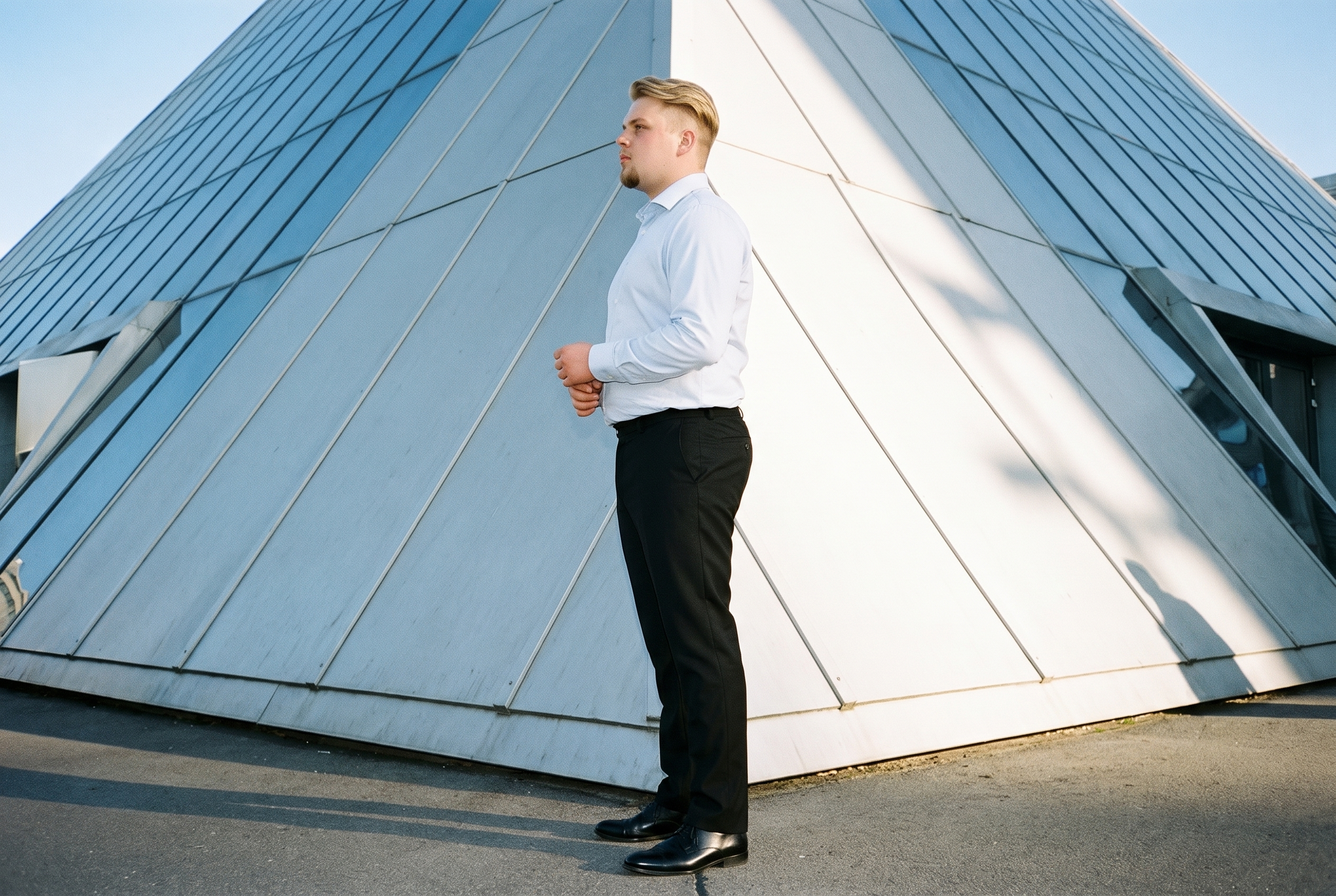 A 19-year-old Caucasian male model wears black tailored trousers in a full-body side view, standing with his hands in front and a natural facial expression. A pyramid building appears as the low-angle backdrop.