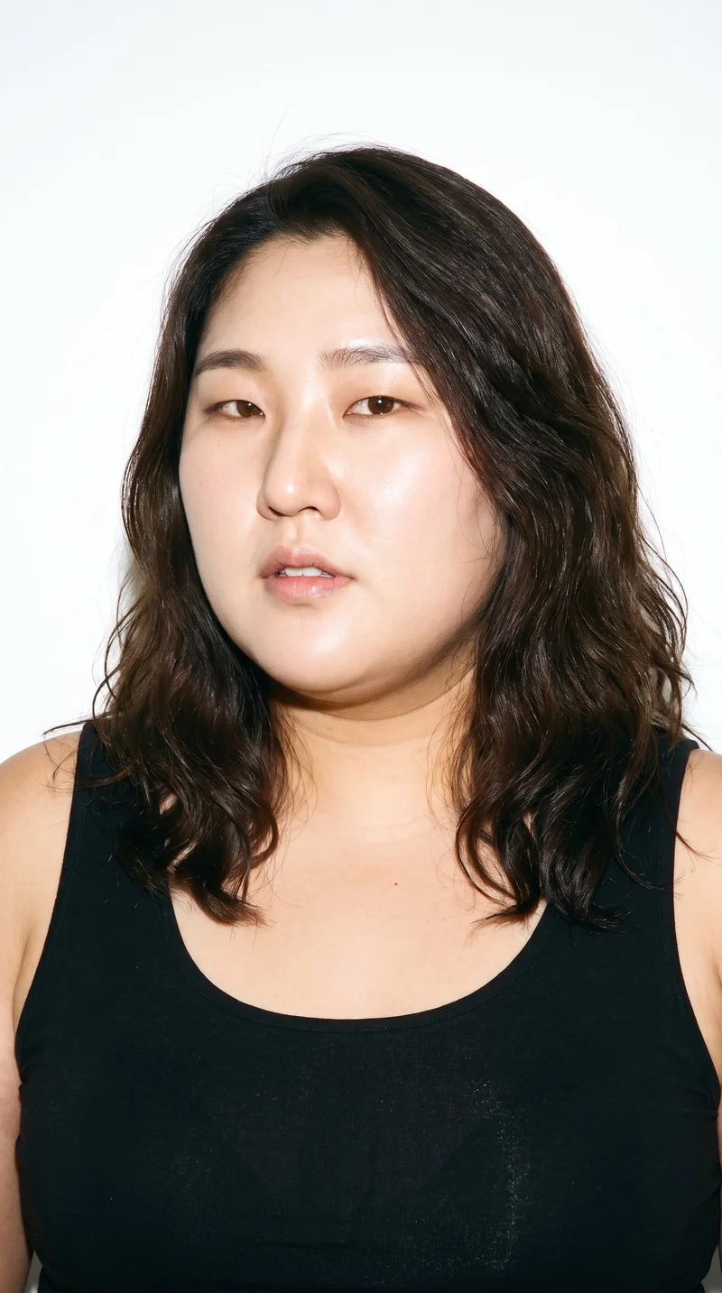 A 37-year-old Korean woman with medium skin and a warm yellow undertone has refined, elongated facial features with high cheekbones. Her eyebrows are medium in thickness, and her eyes are brown. She has full lips and wavy dark-brown hair.