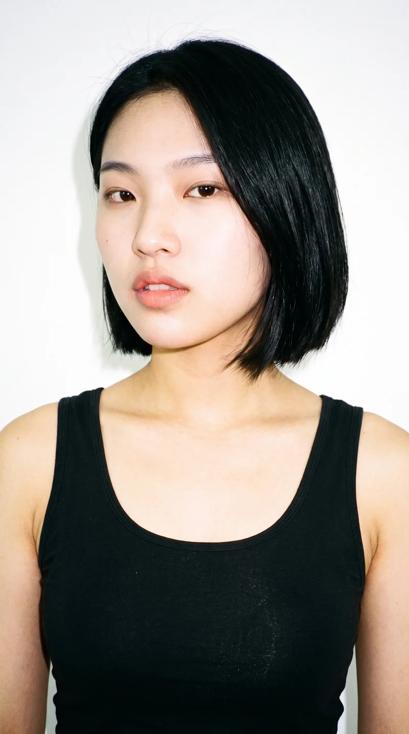 An 18-year-old Chinese woman with fair skin and a warm yellow undertone. Her face has an inverted-triangle shape, with a wider forehead narrowing toward the chin. She has thick eyebrows, brown eyes, and thin lips. Her black hair is cut in an even bob reaching from jaw to chin length.