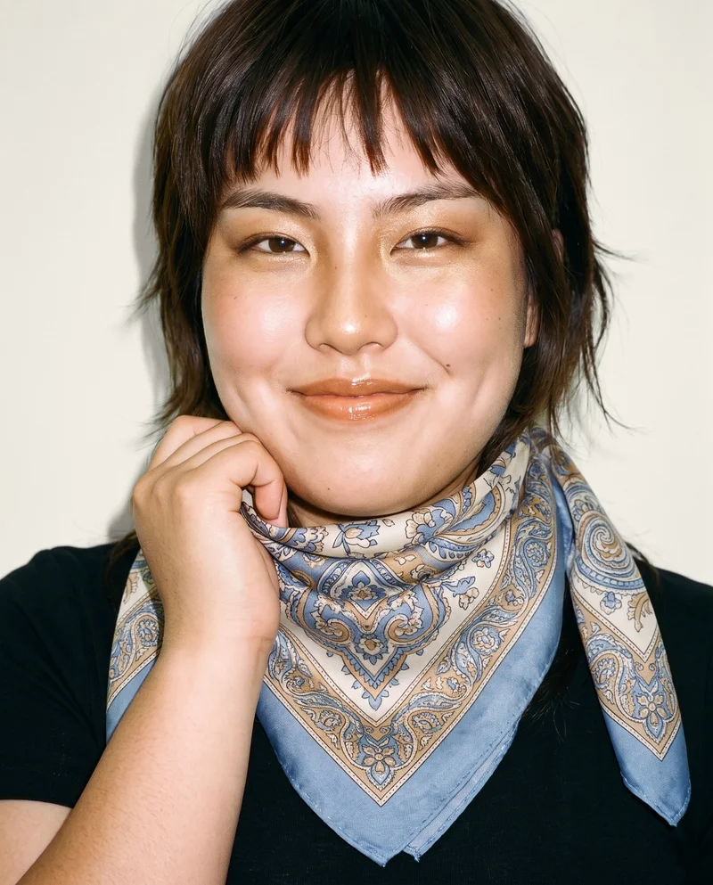A 20-year-old Japanese woman is shown in a front-facing bust view, smiling with her wrist held at her jaw to display a women's patterned silk scarf. An alabaster studio wall forms the backdrop.