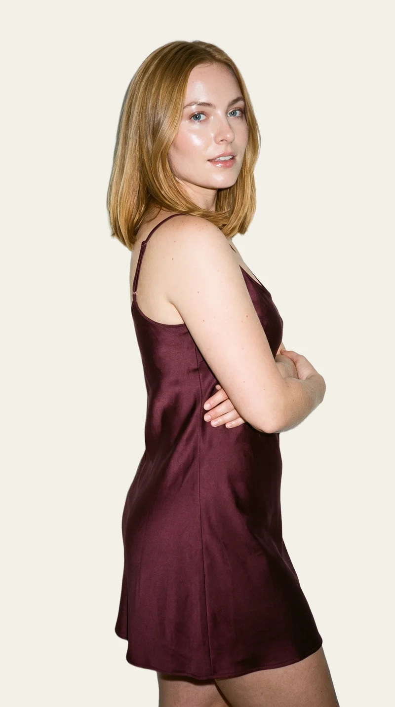 A 25-year-old Caucasian woman wears a burgundy slip dress in a three-quarter-length side view. She is smiling and holding herself in a self-hug pose against an alabaster flat-color backdrop.