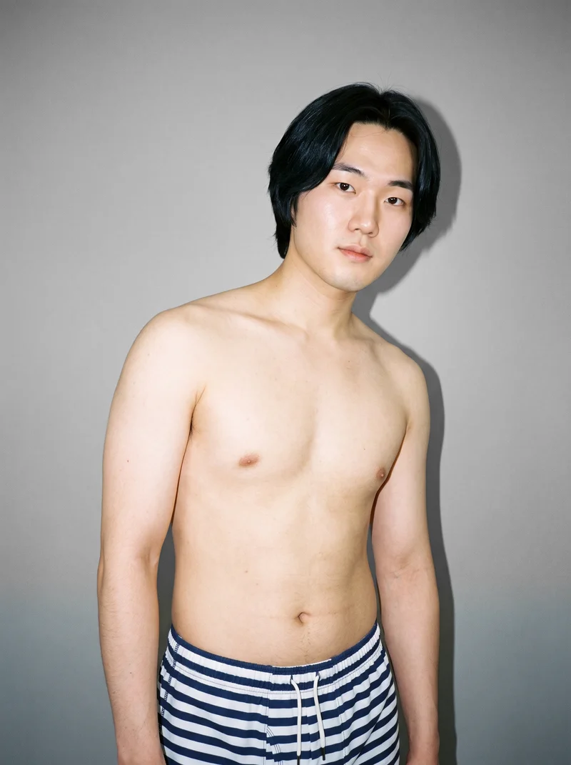 A 25-year-old Korean male model wears striped trunks in an upper-body crop, shown from a front three-quarter angle with a shoulder-drop pose and natural expression. A pebble grey studio-wall backdrop frames the composition.