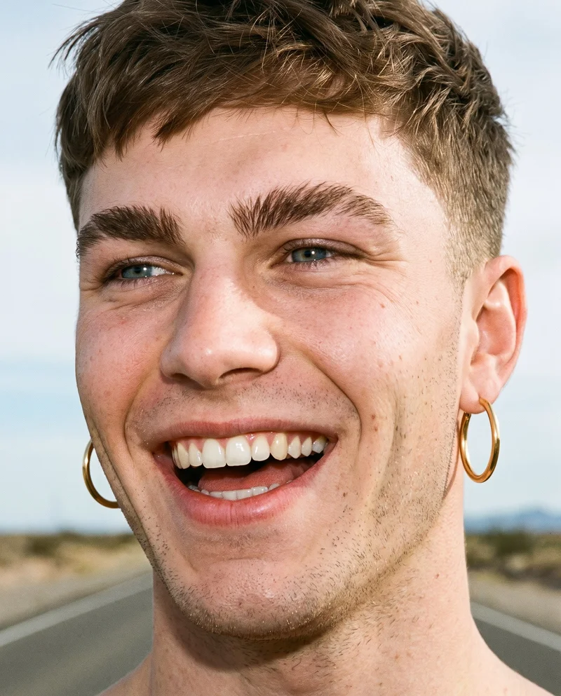 A 20-year-old Caucasian man wears gold hoop earrings in an angled-front head view. His head is level, and he is laughing against a desert highway backdrop.