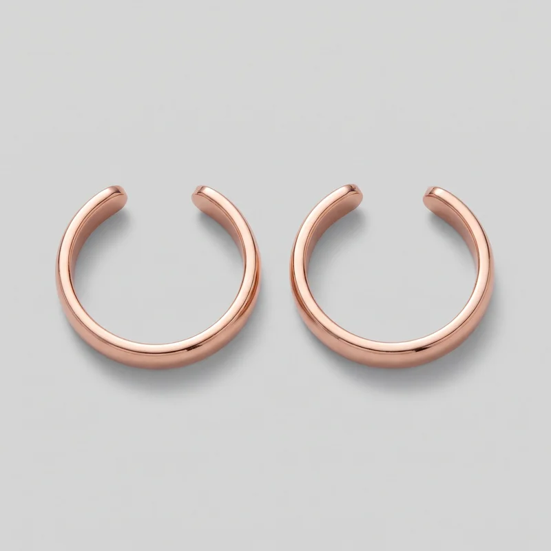 A matching pair of rose gold ear cuffs is shown as standalone jewelry from an angled top view. Each earring has a rounded C-shaped tubular body, smooth domed surfaces, open ends, and a recessed groove along the outer side edge. No posts, hinges, closures, logos, prints, patches, embroidery, or embellishments are visible.