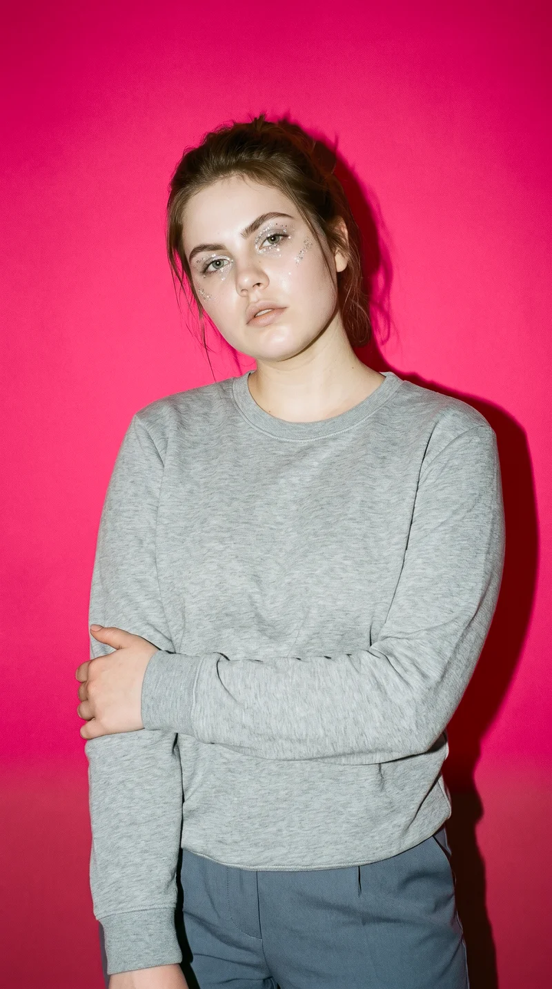 A 20-year-old Slavic woman wears a grey crewneck pullover jumper in an upper-body, front-facing view. She holds one elbow and has a serious expression, with a ruby red studio-wall backdrop.
