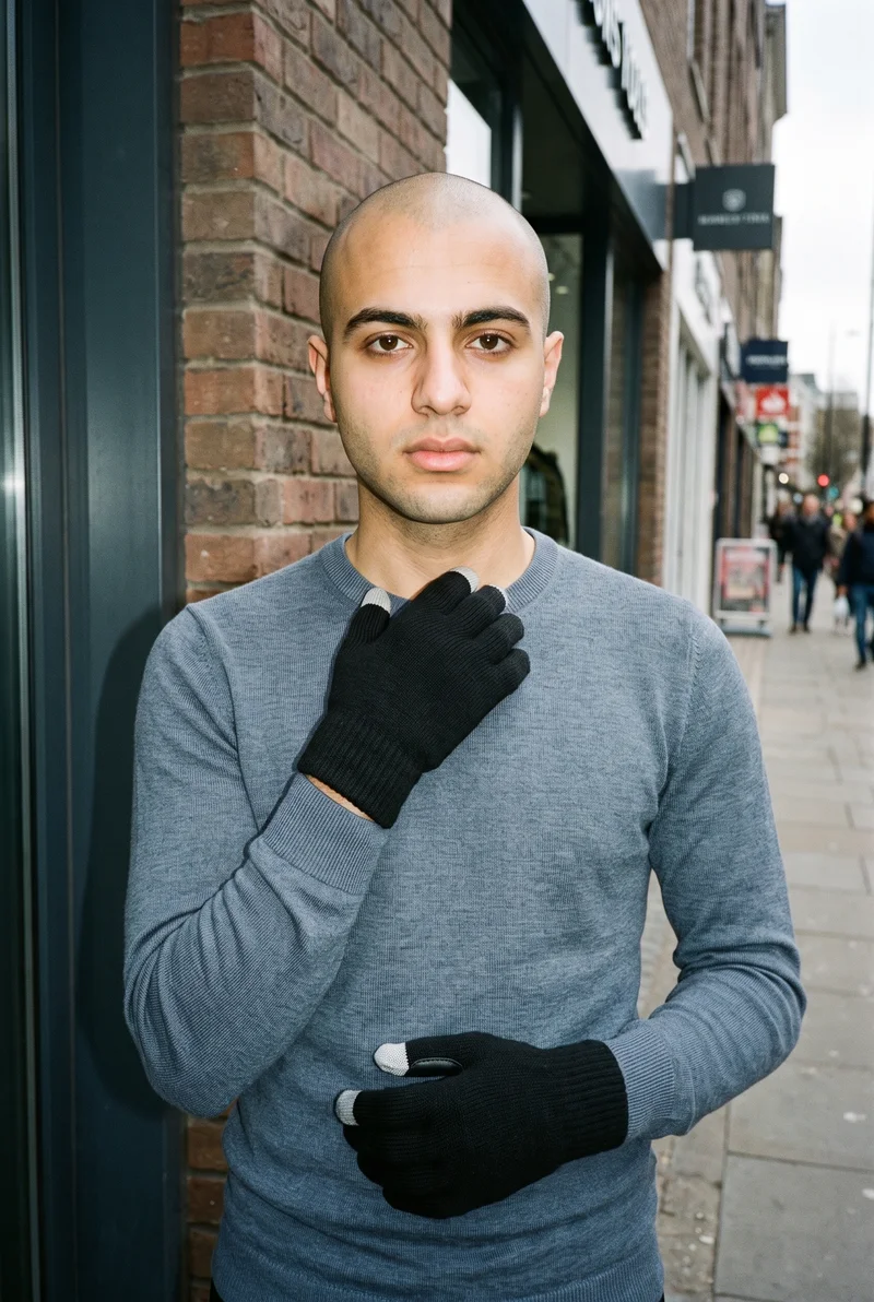 An 18-year-old Arab man is shown from the front in an upper-body view, wearing black touchscreen gloves with one hand held at his collarbone. His serious expression is visible against an urban street backdrop.