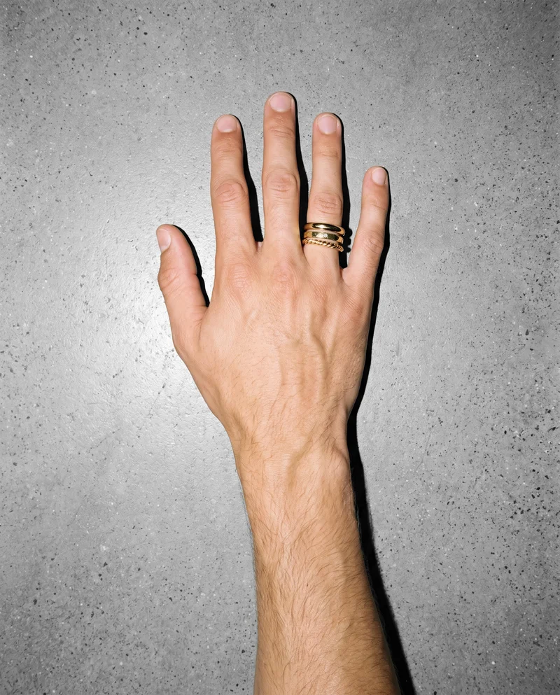 A hand and wrist shown from above at an angle, wearing a men's gold stacking ring set. The Fog Grey studio-wall backdrop frames the close-up composition.