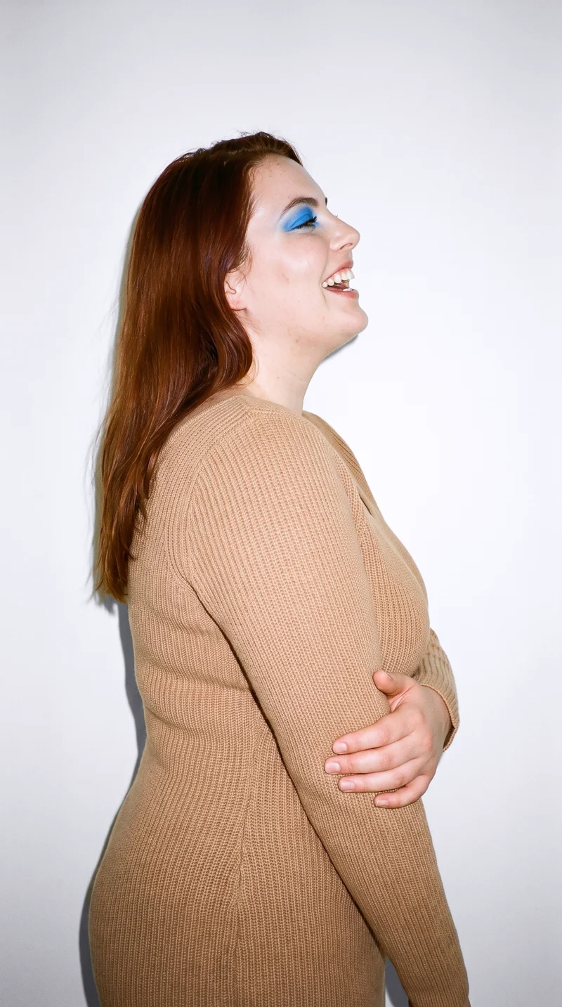 A 23-year-old Caucasian woman wears a camel knit dress in an upper-body side view, holding her elbow and laughing. A porcelain studio-wall backdrop frames the composition.