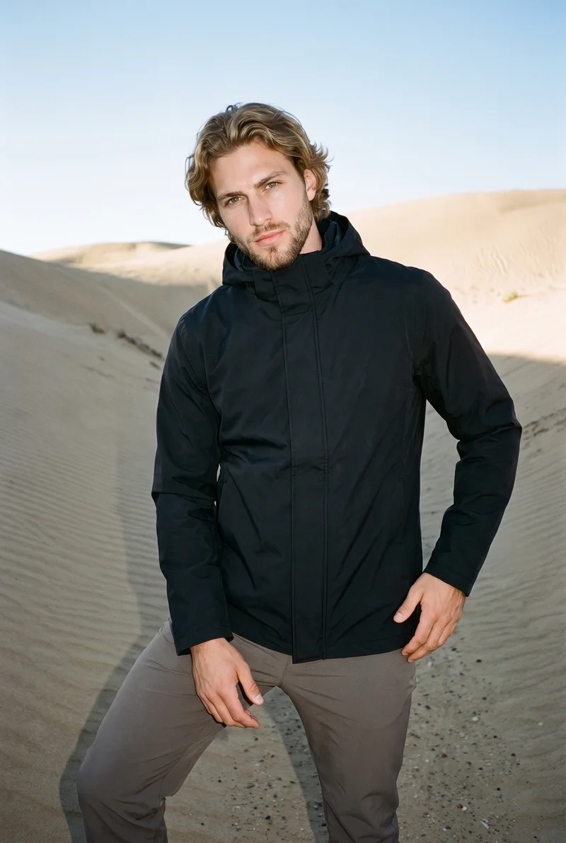 A 24-year-old Caucasian man wears a black hooded raincoat in a three-quarter-length front view, posed with one hip out and a natural expression. Death Valley dunes appear as the backdrop.