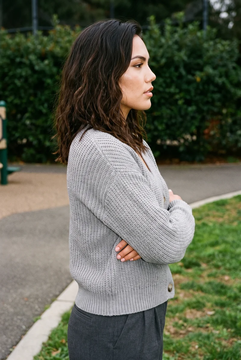 A 24-year-old Indigenous American woman wears a grey knit cardigan in an upper-body side-view composition, with her forearms folded and a serious expression. A playground serves as the backdrop.